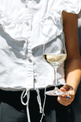 Woman holding a glass of white wine outdoors in sunlight. Stylish casual outfit and relaxed summer mood captured in elegant modern composition.
