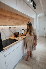A pregnant woman pours coffee into two cups on a bright, modern kitchen countertop with white cabinets and wooden accents, creating a calm, cozy home scene.