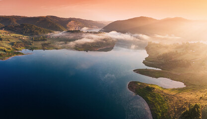 Hills and forests immersing a calm lake with ethereal fog during an orange sunrise, creating a...