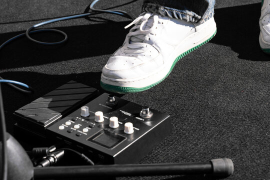 Musician adjusting electric guitar pedalboard during outdoor performance, foot, finger pressing effect pedal and instrument cable, creative live concert stage setup, modern music gear, sound control