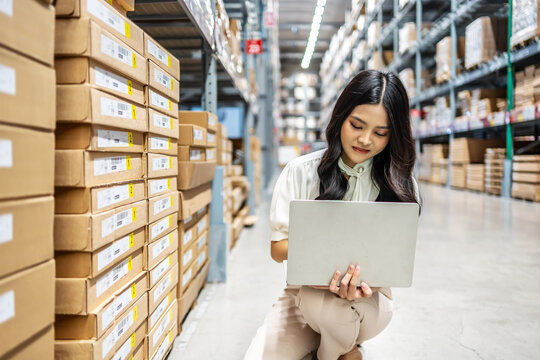 Focused on her task, Young asian woman sitting and holding laptop meticulously updates inventory records, thinking about project while surrounded by shelves filled with various products in a warehouse
