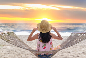 A woman in a straw hat relaxes on a hammock by the beach, enjoying a peaceful sunset over the calm ocean horizon.