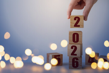 New 2026 Year. Woman stacking cubes with numbers at table, closeup. Bokeh lights