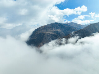 High aerial view of a rugged, dark mountain peak emerging from a thick, swirling layer of white clouds or fog under a bright, patchy blue sky