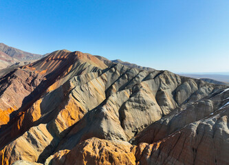 Wide aerial view of a mountain range displaying stunning multicolored, layered sedimentary rock formations and sharp ridges under a clear, bright sky