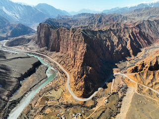 High aerial panorama of a winding road and turquoise river flowing past a colossal, sheer red sandstone cliff formation in a rugged mountain valley