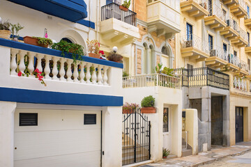 Obraz premium Mediterranean street view showing a house with an iron gate, white garage, terrace with balustrade and flowers, beside a residential building with balconies and wrought-iron railings.