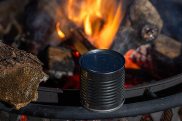 A can of food heating up next to a fire. The fire is raging and the can is silver.