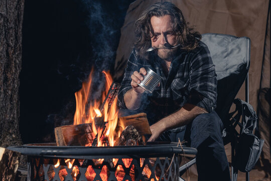 A man camping sitting by the fire eating canned food.