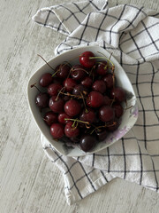 White bowl with dark red cherries on a checkered towel on a wooden surface. Used in food, nutrition, and culinary materials.