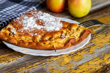A homemade apple cake generously dusted with powdered sugar, presented on a rustic surface. The golden crust, soft texture, and visible apple pieces create a warm and inviting atmosphere, perfect for 