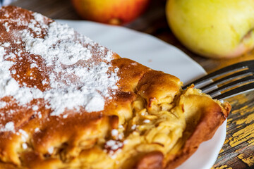 A homemade apple cake generously dusted with powdered sugar, presented on a rustic surface. The golden crust, soft texture, and visible apple pieces create a warm and inviting atmosphere, perfect for 
