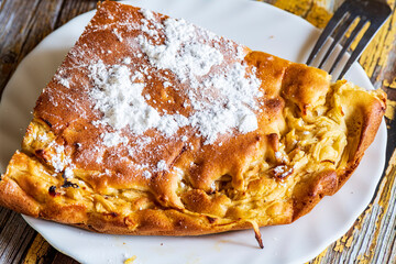 A homemade apple cake generously dusted with powdered sugar, presented on a rustic surface. The golden crust, soft texture, and visible apple pieces create a warm and inviting atmosphere, perfect for 