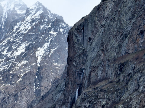 Close-up view of a sheer, textured rock cliff face with a small waterfall, set against a rugged, snow-dusted mountain in the background