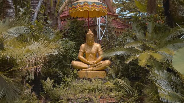A gold colored statue of a meditating Khmer hermit sits under parasol in the temple garden of Wat Svay Romeat Pagoda in Siem Reap, Cambodia. Shot in motion