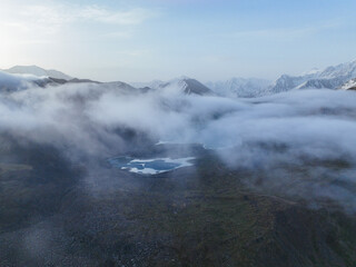 High aerial view of a small mountain lake partially covered by swirling fog and mist, surrounded by rocky slopes and snow-covered peaks at sunrise or dusk