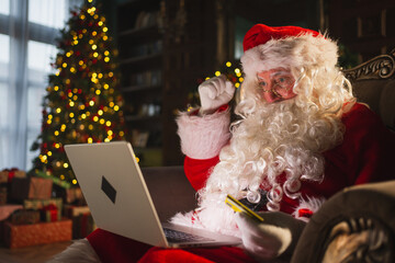 Merry Christmas. Santa Claus shopping online holding laptop paying with gold credit card for...