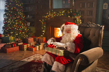 Merry Christmas. Santa Claus sitting in chair packing wrapping gift box in festive living room with...