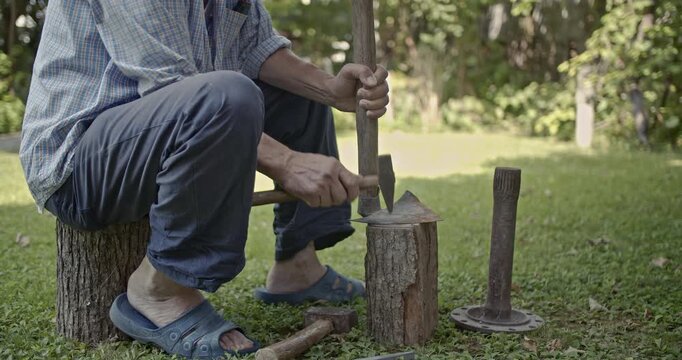 An elderly man meticulously sharpens an axe with a hammer while sitting outdoors on a tree stump. The video captures the traditional craft with clear, authentic sounds of metal striking metal.