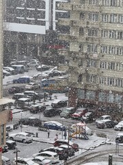 Snowfall over city buildings, cars, and a small playground. Used in urban, architecture, and climate-related materials.