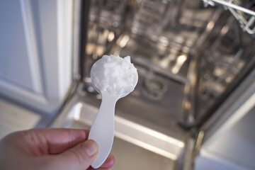 Hand adding dishwasher salt with white plastic spoon to open appliance in kitchen.