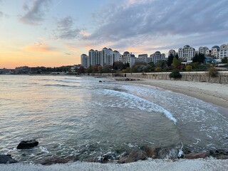 City beach at sunset with calm waves and modern buildings on the coast. Peaceful seaside evening with soft clouds and warm light.