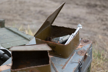 An open, rusty ammunition can is filled with spent casings, belt links, and an RPG-7 tail fin stabilizer. Discarded on a battlefield, these remnants serve as a stark artifact of recent combat.