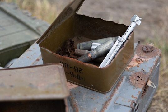 An open, rusty ammunition can is filled with spent casings, belt links, and an RPG-7 tail fin stabilizer. Discarded on a battlefield, these remnants serve as a stark artifact of recent combat.