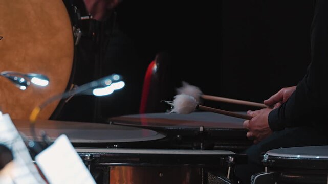 A close-up view of a percussionist's hand playing mallets on a timpani. The background features a large bass drum and sheet music, creating an musical atmosphere