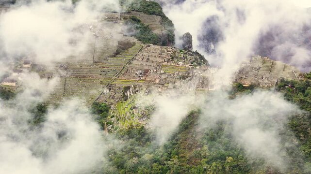 Aerial circles high above the clouds, focusing on the Machu Picchu citadel below. The shot clearly shows lines of tourists walking in single file on the different levels of the iconic green terraces