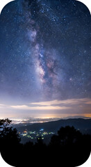 Naklejka premium The stunning milky way galaxy arches over a distant, glowing cityscape at night, with silhouetted trees in the foreground