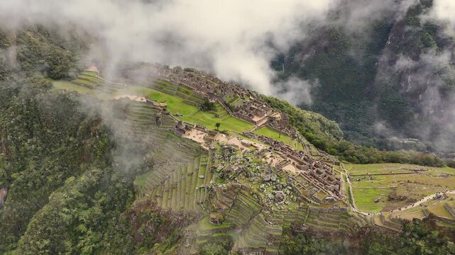 Breathtaking drone shot approaches Machu Picchu from a distance, flying towards the ancient citadel. As clouds pass by, the camera elegantly pans downward to reveal a close-up of the intricate stone