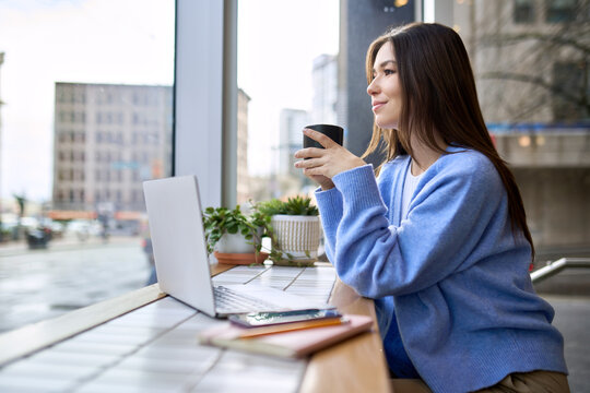 Happy calm lady student enjoying warm hot drink with mug in hands working or studying online. Young smiling woman sitting holding cup drinking tea or coffee relaxing in cafe looking through window