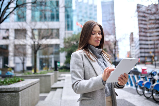Young busy business woman professional or female student standing on big city street using digital tablet fintech pad device technology looking at tab thinking of financial success.