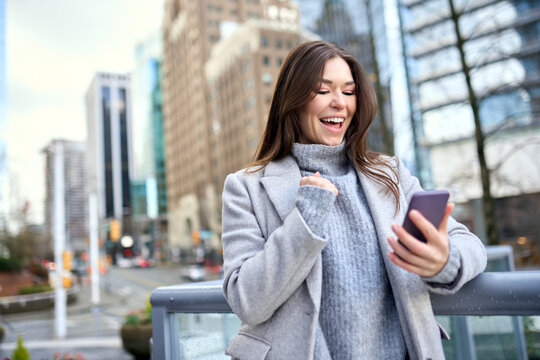 Happy young woman excited winner holding cellphone using mobile phone winning online, receiving great news sms offer looking at smartphone, getting new job celebrating success standing on city street. - Powered by Adobe