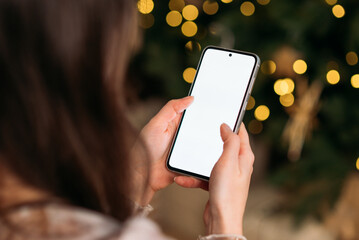 Close-up of woman using smartphone with blank white screen near Christmas tree with golden festive lights.