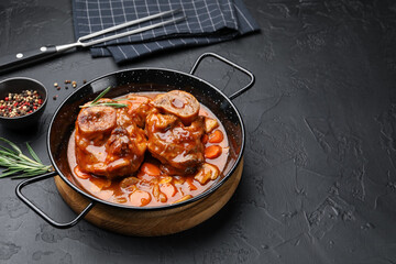Tasty beef ossobuco with carrot, mushrooms and rosemary in frying pan on dark textured table, closeup