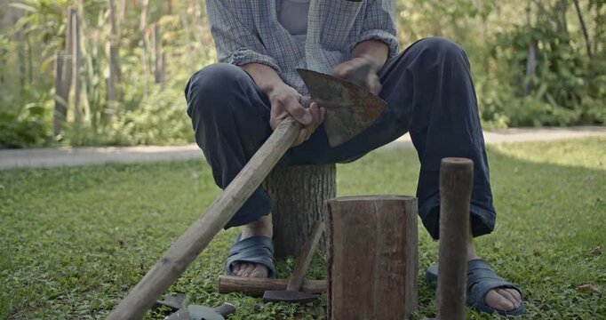 Watch a man meticulously sharpen an old, rusty axe by hand with a stone, sitting on a log in a green outdoor setting. This video features the authentic sounds of his traditional craftsmanship.