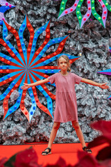 A teenage girl stands confidently in front of a large street Christmas tree adorned with bright, colorful decorations. The festive atmosphere is warm and inviting. 