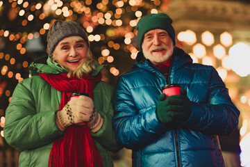 Funny warm moment outdoors with a couple of grandparents smiling holding cups in festive city lights and winter coats