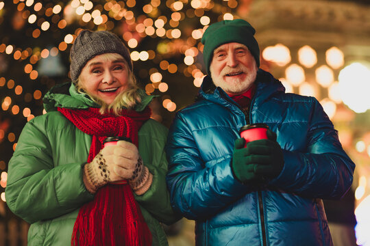Funny warm moment outdoors with a couple of grandparents smiling holding cups in festive city lights and winter coats - Powered by Adobe