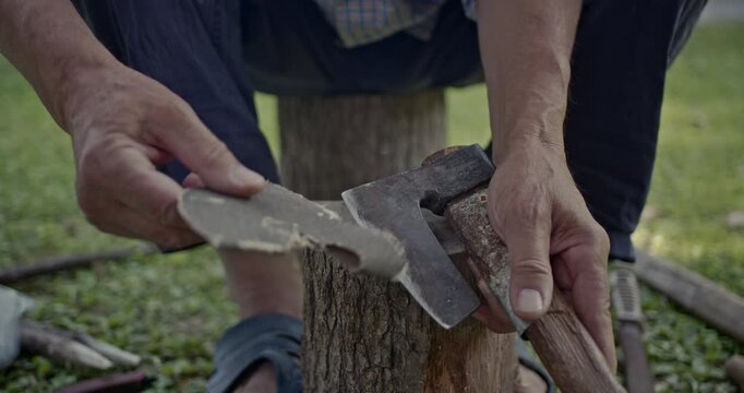 A close-up view of hands diligently sharpening an axe blade with sandpaper on a tree stump outdoors. The authentic sounds enhance the natural and focused process of tool maintenance.