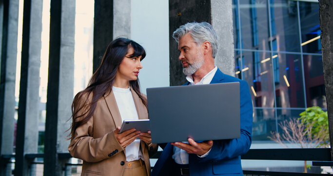 Adult Caucasian businessman in formal suit holding a laptop, collaborating with elegant woman in beige blazer holding a tablet.