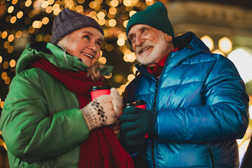Joyful senior couple outdoors sharing warm drinks under festive Christmas lights in a snowy city celebration