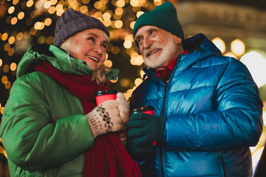 Joyful senior couple outdoors sharing warm drinks under festive Christmas lights in a snowy city celebration