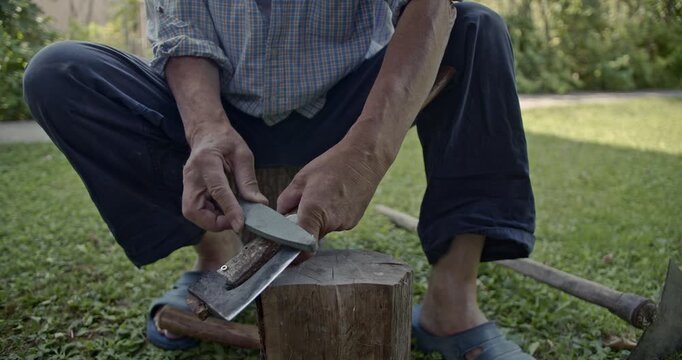 An elderly man meticulously sharpens an axe blade using a whetstone while sitting outdoors. This video features clear, authentic sounds of the traditional sharpening process.