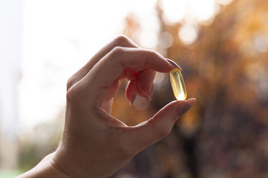 Closeup of a person holding a vitamin D 3 capsule against colourful trees in fall.