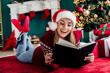 Funny Christmas moment as girl in a Santa hat reads a book at home beside a twinkling tree and...