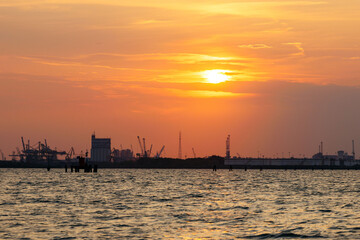 Fototapeta premium Venice, Italy - June 23, 2025: Sunset over industrial harbor with silhouettes of cranes and buildings reflecting on calm water, creating a serene yet dynamic atmosphere at dusk
