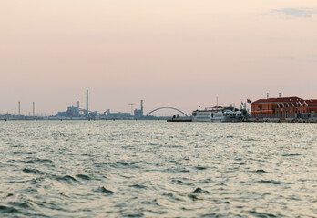Venice, Italy - June 23, 2025: Scenic view of industrial waterfront with calm water reflecting pastel sky, featuring boats and buildings along the shoreline at dusk